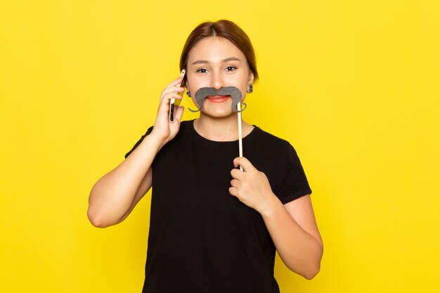 Uma jovem mulher de vestido preto, vista frontal, posando com bigode postiço e falando ao telefone, sorrindo em amarelo