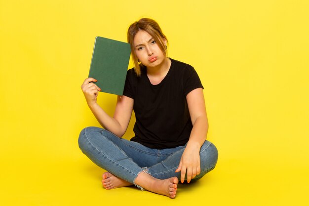 Uma jovem mulher sentada de frente, de camisa preta e calça jeans, segurando um caderno verde, pensando deprimido no amarelo