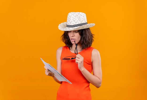 Uma jovem pensativa, com cabelo curto e uma camisa laranja com chapéu de sol, olhando para um mapa
