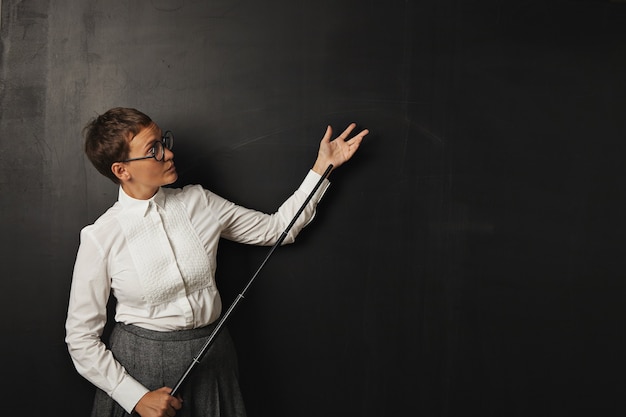 Uma jovem professora branca, séria, com blusa de botão e saia de tweed parada em um quadro negro segurando um ponteiro