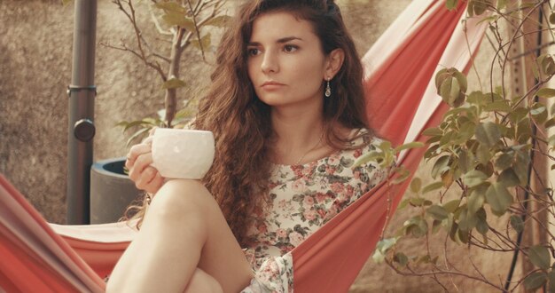 Uma jovem ruiva de vestido floral lê um livro em uma rede em um pequeno jardim urbano