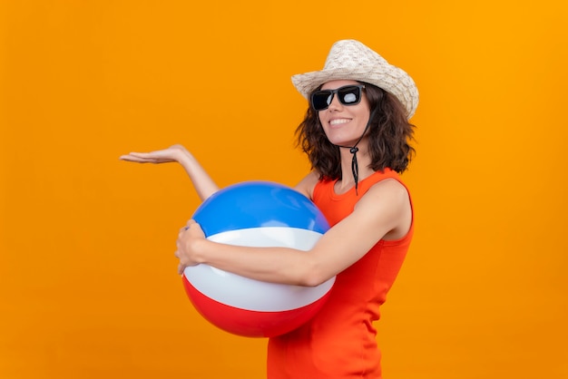 Uma jovem satisfeita com cabelo curto e uma camisa laranja usando chapéu de sol e óculos escuros segurando uma bola colorida inflável