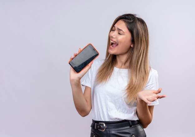 Uma jovem séria em uma camiseta branca cantando uma música segurando o celular como um microfone em uma parede branca