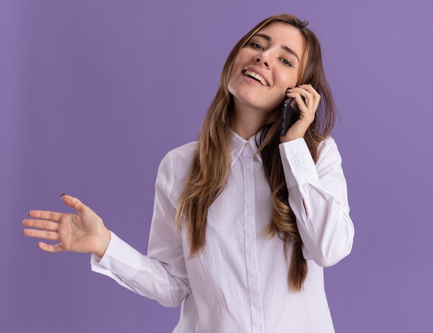 Uma jovem sorridente e bonita caucasiana com a mão aberta e fala no telefone, isolada na parede roxa com espaço de cópia