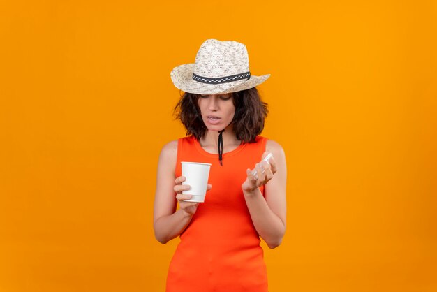 Uma jovem surpresa com cabelo curto e uma camisa laranja com chapéu de sol abrindo um copo de plástico