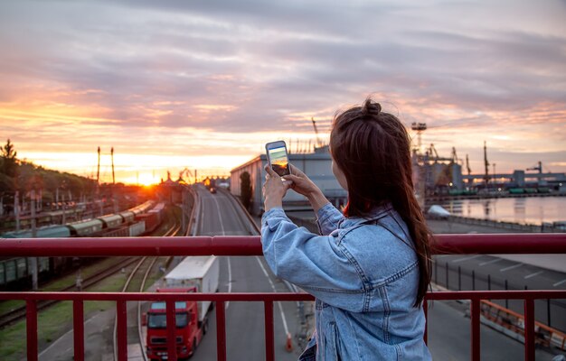 Uma jovem tira uma foto de um belo pôr do sol de uma ponte em seu telefone.