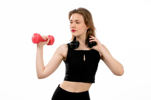 Uma jovem vista frontal na camisa preta e calça preta, fazendo esporte com halteres vermelhos no branco