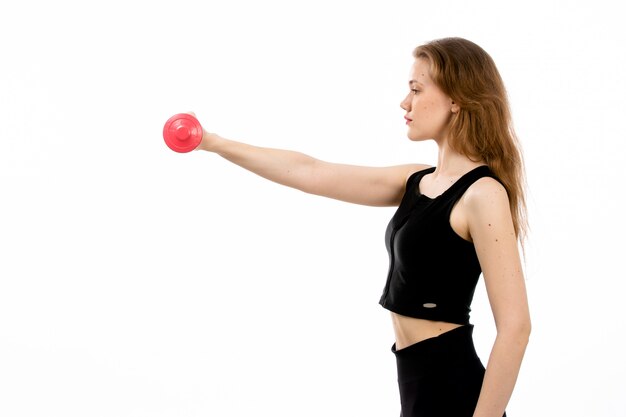 Uma jovem vista frontal na camisa preta e calça preta, fazendo esporte com halteres vermelhos no branco