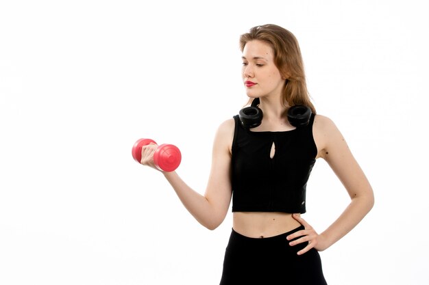 Uma jovem vista frontal na camisa preta e calça preta, fazendo esporte com halteres vermelhos no branco