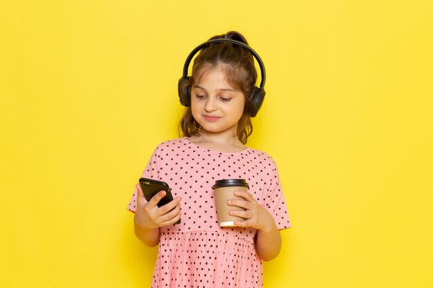 Uma linda criança de vestido rosa segurando e usando o telefone, ouvindo música com café na mesa amarela