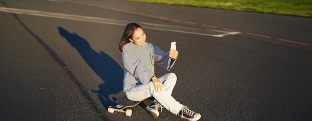Foto grátis uma linda garota coreana tira uma selfie no smartphone tira uma foto com seu skate no dia ensolarado