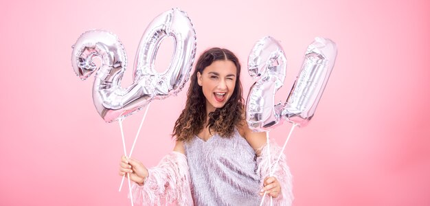 Uma linda garota em um clima festivo pisca em um fundo rosa do estúdio e segura em suas mãos balões de prata para o conceito de ano novo