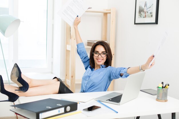 Uma linda garota morena, sentada à mesa no escritório, as pernas cruzadas sobre a mesa. Ela usa camisa azul e saia preta com sapatos. Ela parece muito enérgica.