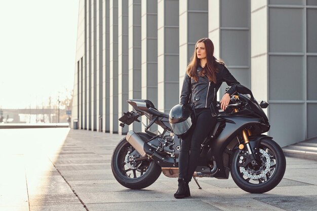 Uma linda garota motociclista vestindo jaqueta de couro preta encostada em sua superbike do lado de fora de um prédio em um dia ensolarado.