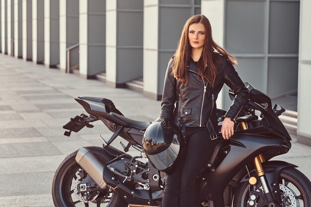 Uma linda garota motociclista vestindo jaqueta de couro preta encostada em sua superbike do lado de fora de um prédio.