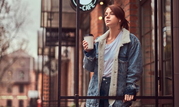 Uma linda garota tatuada vestindo um casaco jeans segurando a xícara com café para viagem do lado de fora perto do café.