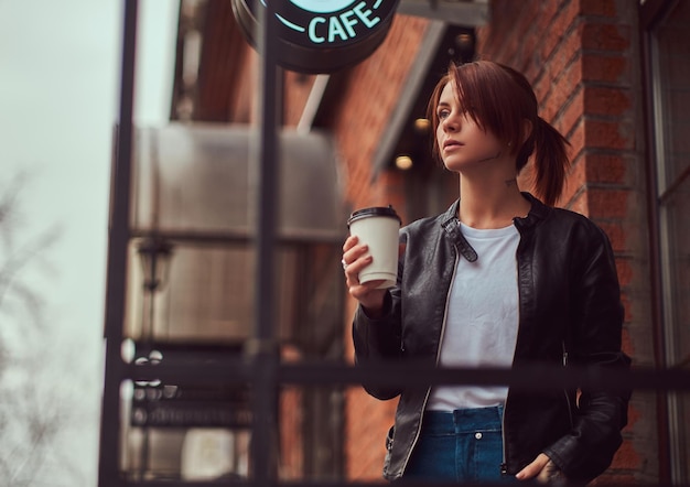 Uma linda garota vestindo uma jaqueta de couro com uma mochila segurando a xícara com café para viagem do lado de fora perto do café.