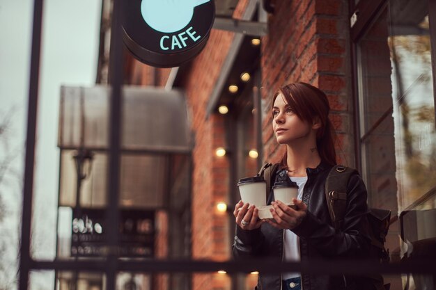 Uma linda garota vestindo uma jaqueta de couro com uma mochila segurando xícaras com café para viagem do lado de fora perto do café.