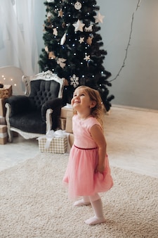Uma linda garotinha com um lindo vestido se diverte muito perto da árvore de natal em casa
