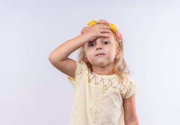 Uma linda garotinha vestindo uma camisa amarela com uma faixa floral, mantendo as mãos na cabeça em uma parede branca