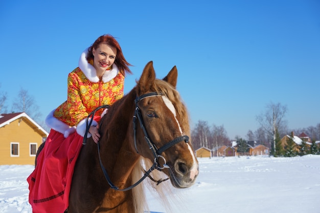 Uma linda jovem com uma jaqueta vermelho-amarela quente monta um cavalo em um dia gelado e ensolarado de inverno. Pratica esportes equestres na temporada de inverno
