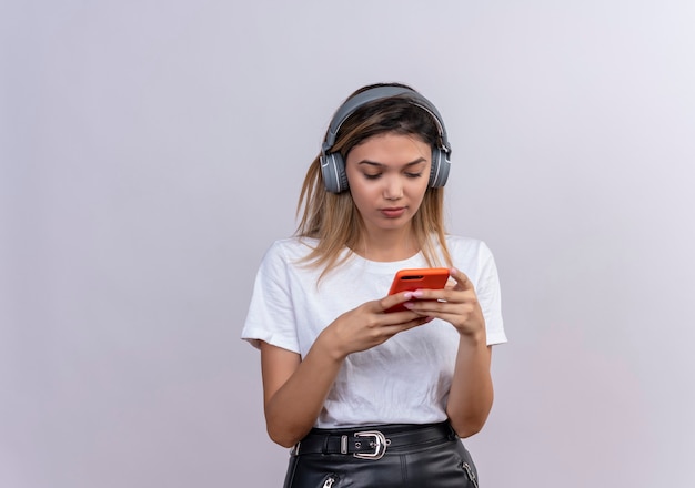 Uma linda jovem em uma camiseta branca com fones de ouvido olhando para o celular e tocando-o em uma parede branca