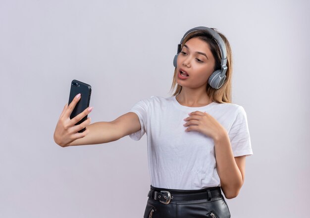 Uma linda jovem em uma camiseta branca usando fones de ouvido tirando uma selfie com o smartphone em uma parede branca