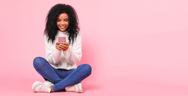 Uma linda mulher de etnia africana está sentada no chão em posição de lótus, olhando para a tela de um smartphone nas mãos, rindo de alegria