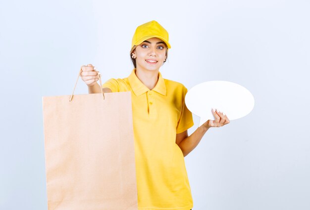 Uma linda mulher de uniforme amarelo segurando um saco de papel artesanal em branco marrom e um balão de fala em branco.