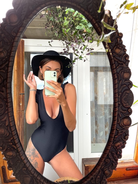 Uma linda mulher em casa tira foto de selfie no espelho do celular para histórias e postagens nas redes sociais, usando maiô preto de verão