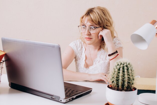 Uma linda mulher em uma blusa branca está sentada no escritório e olha surpresa para o monitor do laptop