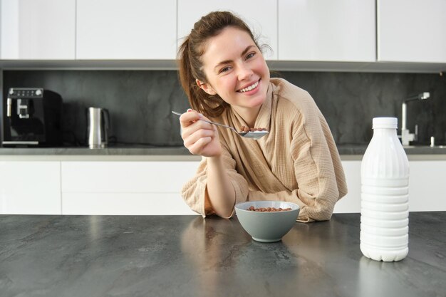 Uma linda mulher rindo de roupão come cereais com leite segurando uma colher sorrindo e parecendo feliz