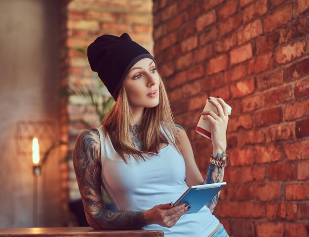 Uma loira sexy tatuada em uma t-shirt e um chapéu com uma xícara de café e computador tablet em uma sala com interior loft.