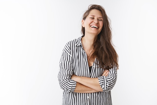 Uma meia-idade feliz rindo sinceramente fecha os olhos e cruza as mãos sobre o peito se divertindo com um lindo presente feito pelas crianças, rindo alegremente enquanto posam com uma blusa listrada sobre a parede branca