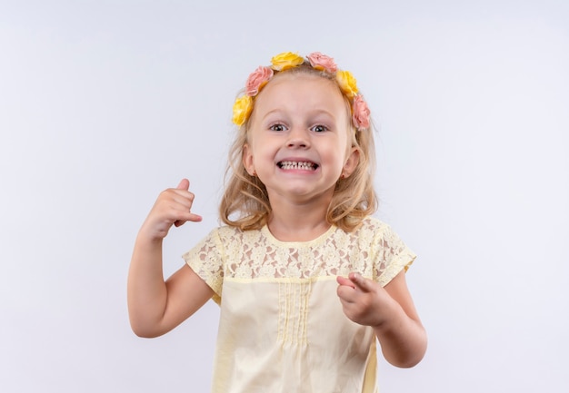 Uma menina alegre e fofa vestindo uma camisa amarela com uma faixa floral, mostrando um gesto de me ligar enquanto aponta em uma parede branca