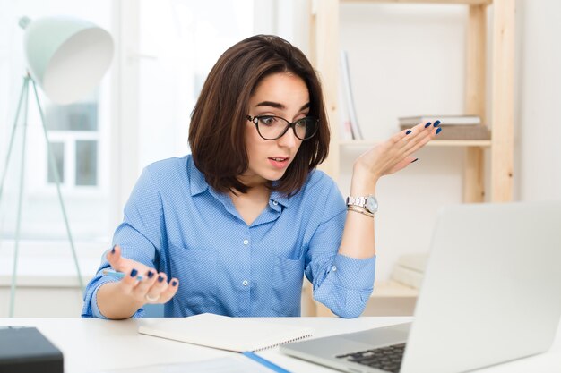 Uma menina bonita morena está sentada à mesa no escritório. Ela usa camisa azul e óculos pretos. Ela está perdida com o trabalho.