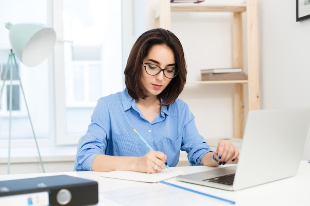 Uma menina bonita morena está trabalhando na mesa no escritório. Ela usa camisa azul e óculos pretos. Ela está escrevendo a sério.