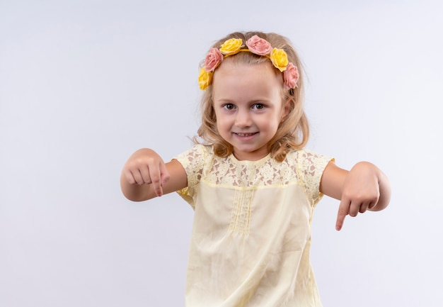 Uma menina bonita vestindo uma camisa amarela com uma faixa floral apontando para baixo com os dedos indicadores em uma parede branca