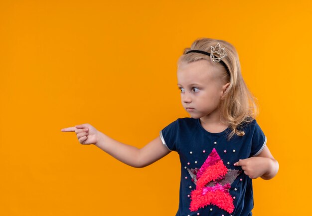 Uma menina bonita vestindo uma camisa azul marinho com uma faixa na cabeça apontando com o dedo indicador enquanto olha para o lado em uma parede laranja