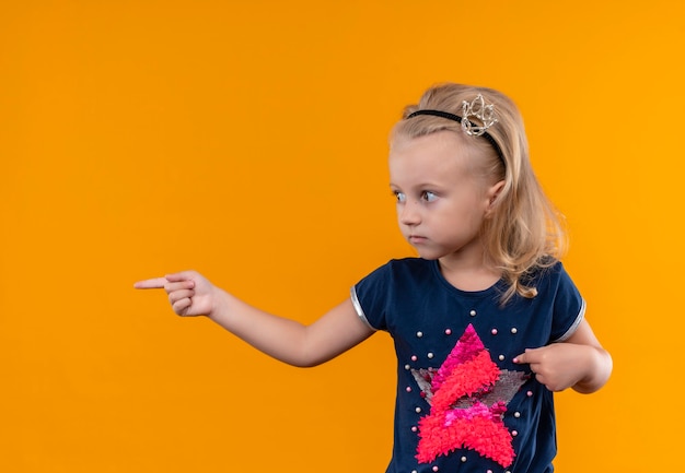 Uma menina bonita vestindo uma camisa azul marinho com uma faixa na cabeça apontando com o dedo indicador enquanto olha para o lado em uma parede laranja