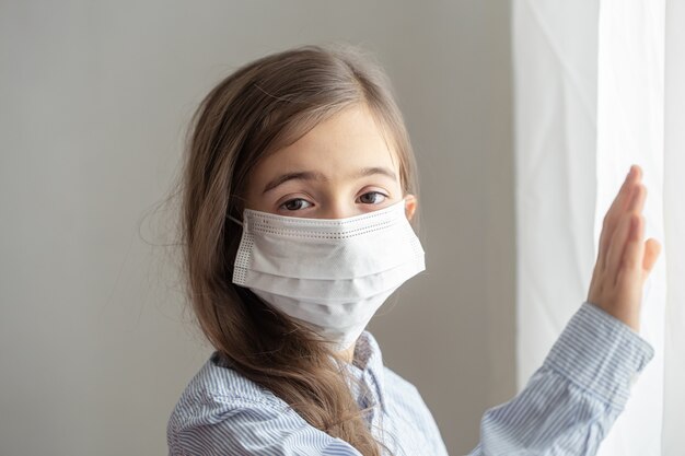 Uma menina bonitinha com uma máscara protetora descartável do coronavírus. Conceito de infância durante a pandemia e a quarentena.