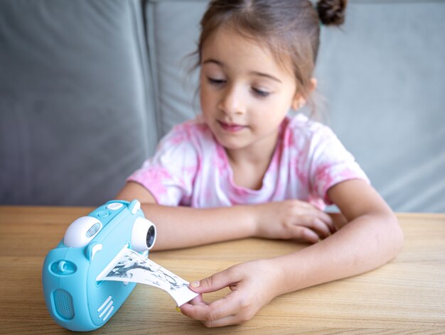 Uma menina bonitinha olha para a câmera de brinquedo azul de seus filhos para impressão instantânea de fotos.