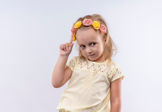 Uma menina bonitinha vestindo uma camisa amarela com uma faixa floral apontando para cima com o dedo indicador em uma parede branca