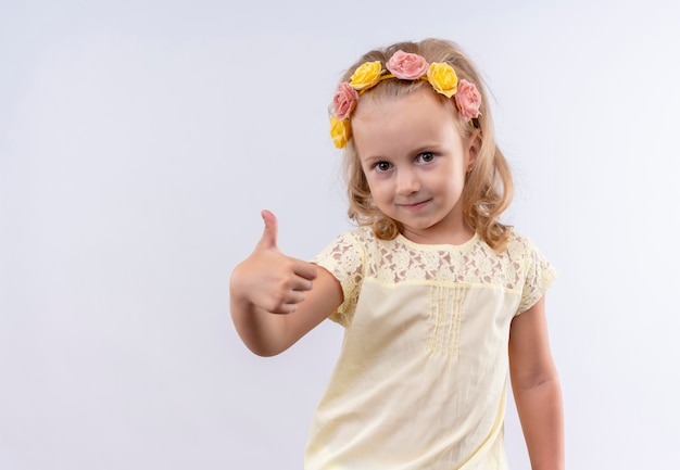 Uma menina bonitinha vestindo uma camisa amarela com uma faixa floral mostrando os polegares para cima enquanto olha para uma parede branca