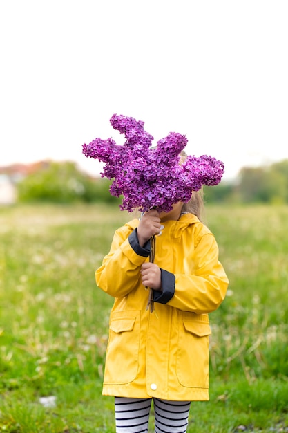 Uma menina com uma jaqueta amarela cobre o rosto com um buquê de lilases. Criativo