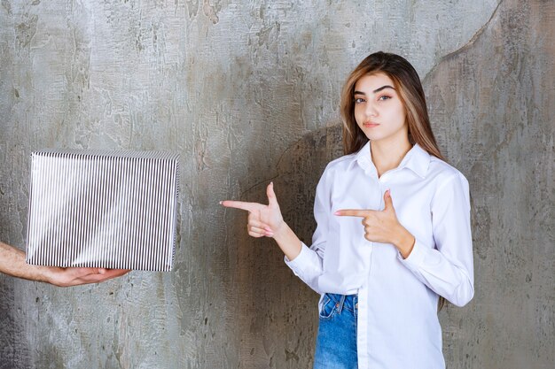 Uma menina de camisa branca em pé em uma parede de concreto está recebendo uma caixa de presente prateada e parece atenciosa