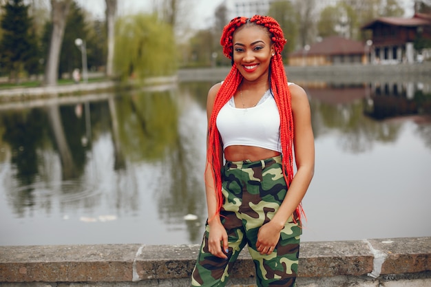 Uma menina de pele escura nova e elegante com dreads vermelho que anda no parque do verão perto do rio