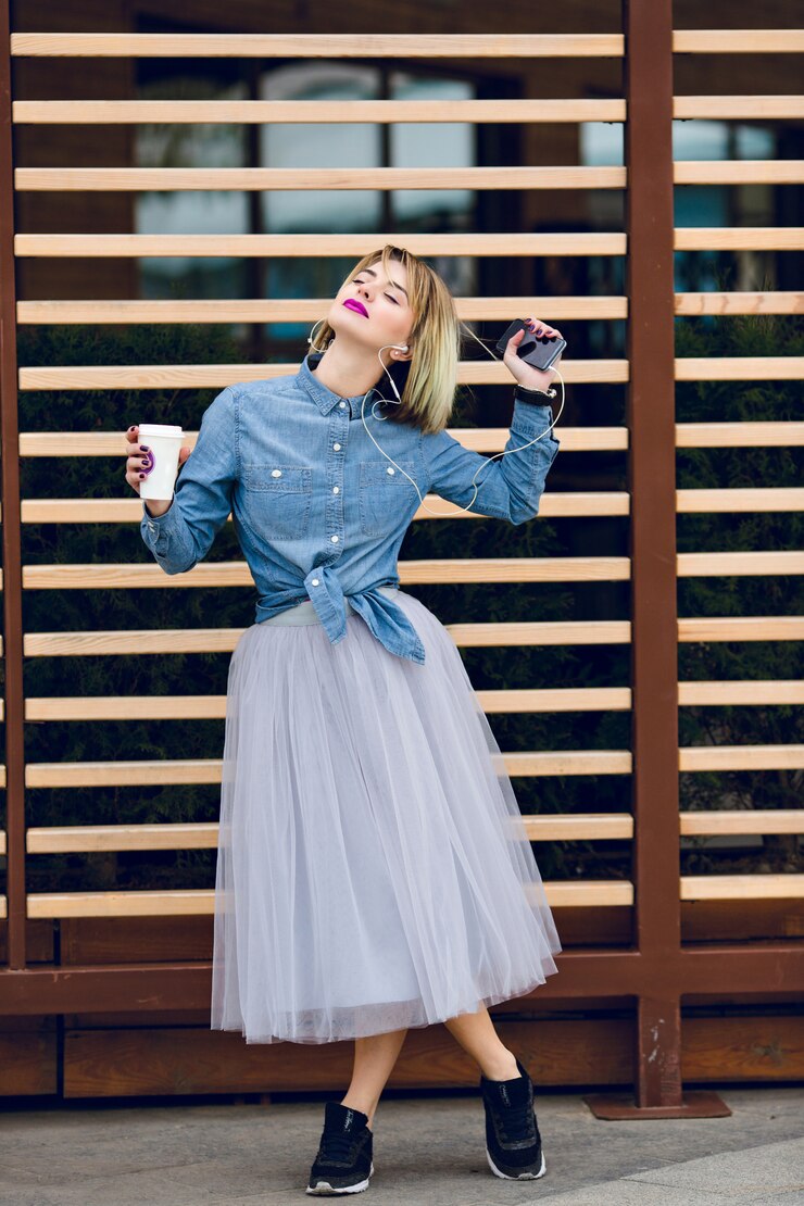 Foto grátis uma menina loira de pé com lábios rosados, segurando uma xícara de café e ouvindo música em um smartphone com madeira listrada se recusa atrás