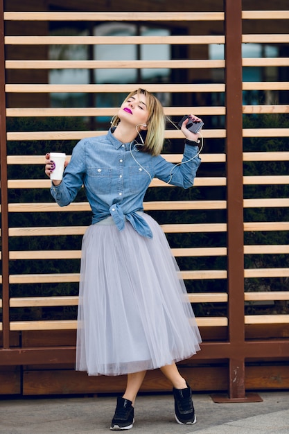 Uma menina loira de pé com lábios rosados, segurando uma xícara de café e ouvindo música em um smartphone com madeira listrada se recusa atrás