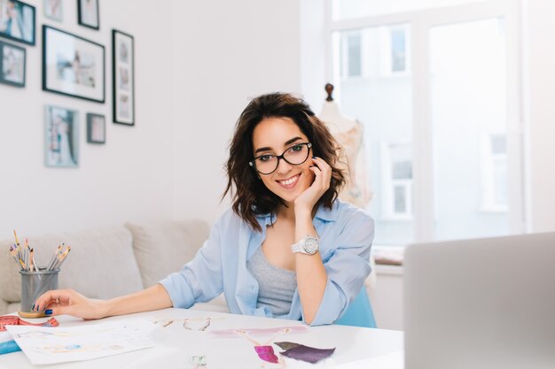 Uma menina morena sorridente com uma camisa azul está sentada à mesa na oficina. Ela tem cetches e lápis na mesa. Ela está sorrindo para a câmera.